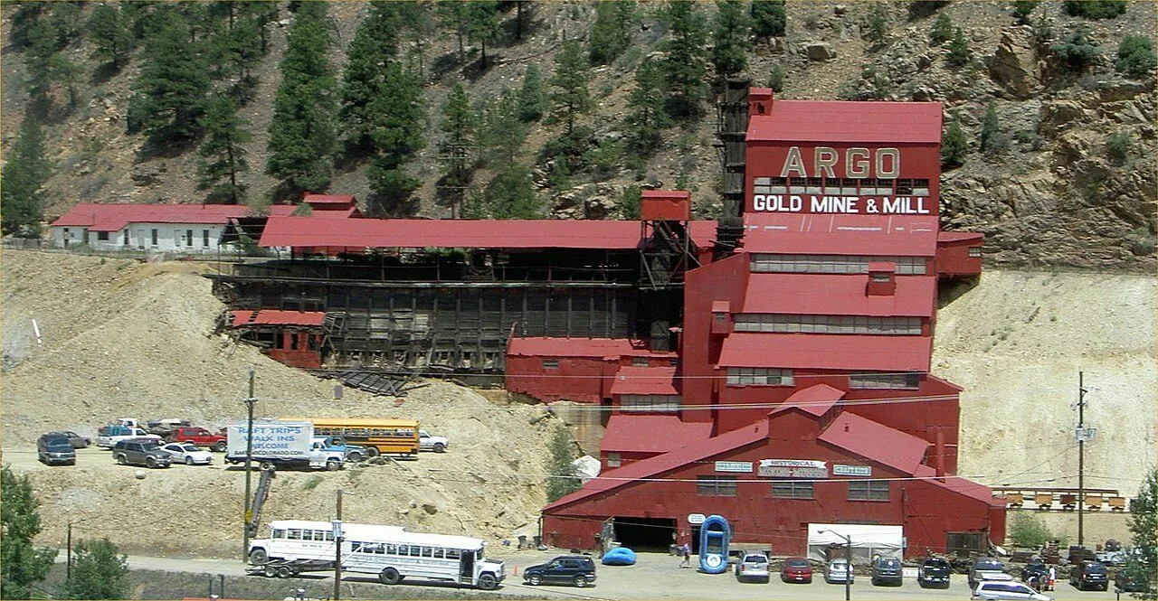 The red Argo Gold Mine & Mill stepping up the hillside above Riverside Drive, ARGO GOLD MINE & MILL painted on the upper mill, with buses and cars parked below, in June 2006