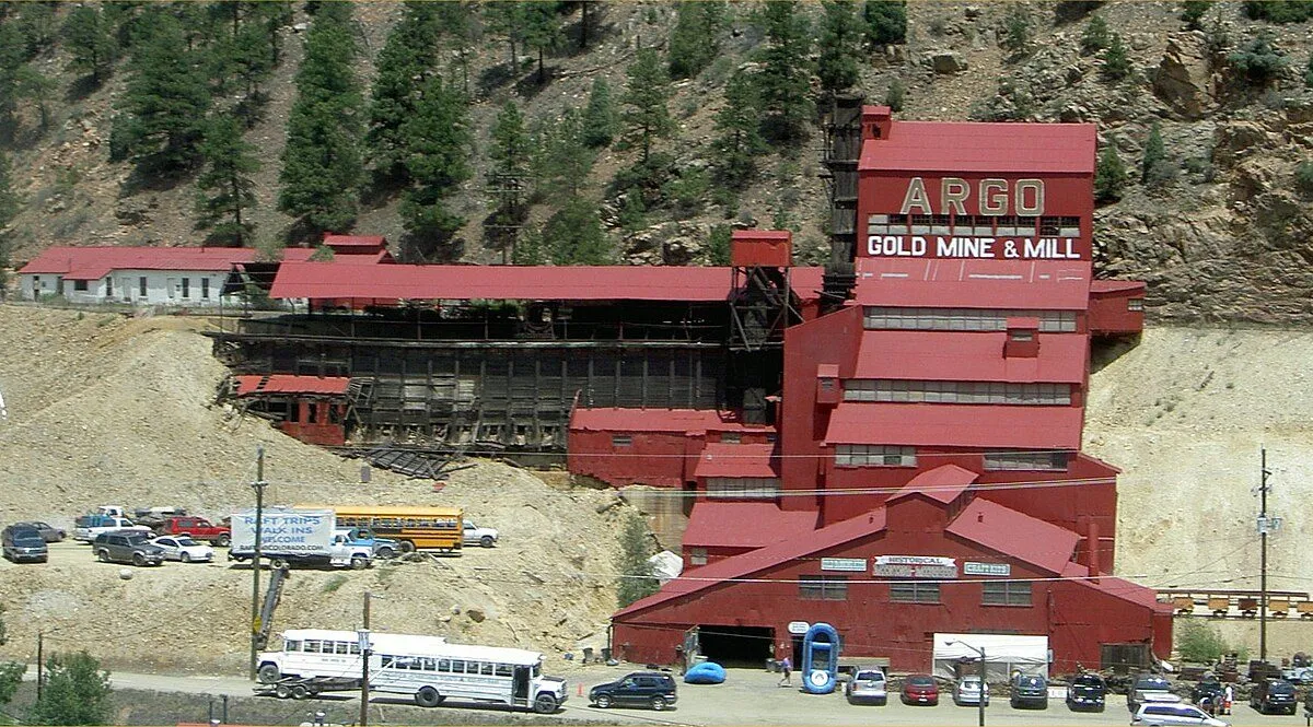 The red Argo Gold Mine & Mill stepping up the hillside above Riverside Drive, ARGO GOLD MINE & MILL painted on the upper mill, with buses and cars parked below, in June 2006