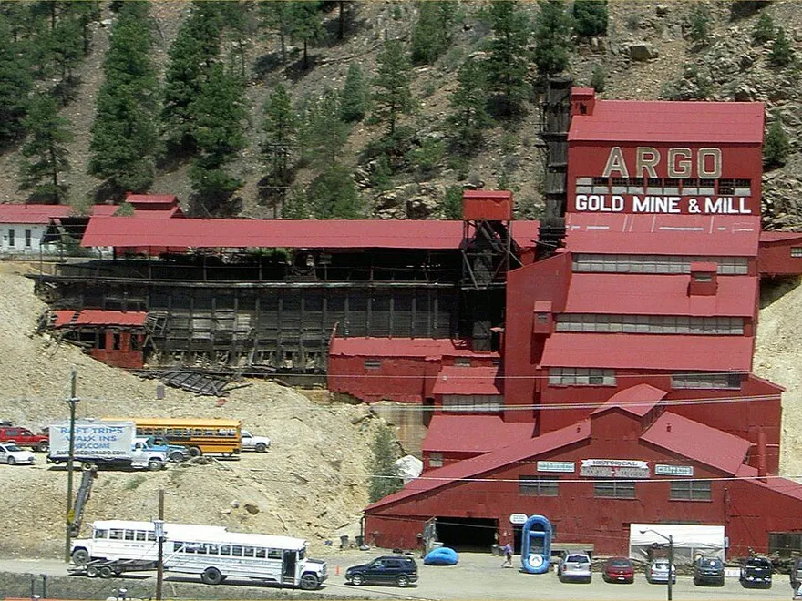 The red Argo Gold Mine & Mill stepping up the hillside above Riverside Drive, ARGO GOLD MINE & MILL painted on the upper mill, with buses and cars parked below, in June 2006